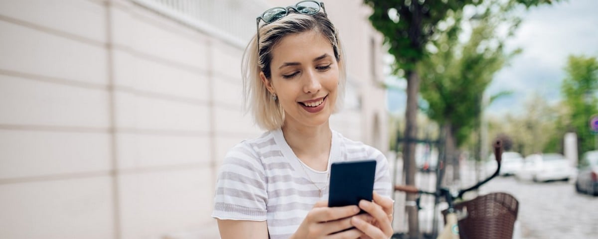 Jeune femme souriante et heureuse, aux cheveux blonds, assise sur un banc, utilisant un téléphone intelligent.